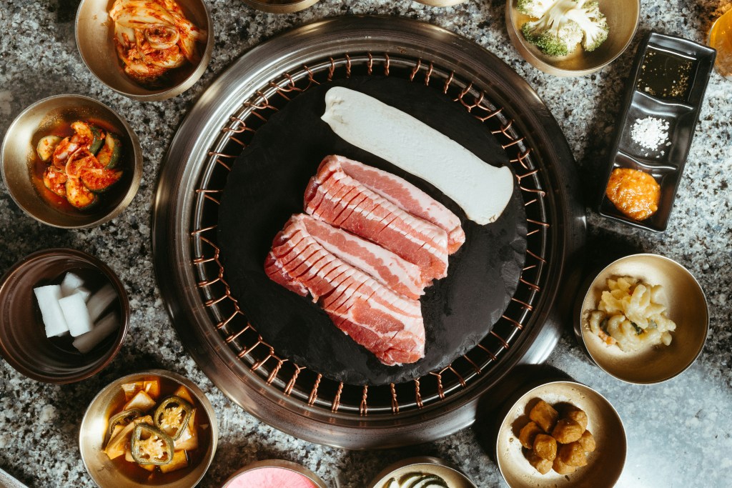Raw sliced pork belly and a king oyster mushroom on a round Korean BBQ grill surrounded by banchan